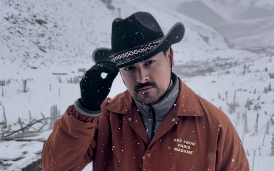 Kevin in a snowy mountain valley, tipping a black cowboy hat, in a rust jacket that reads Not from Paris, Madame